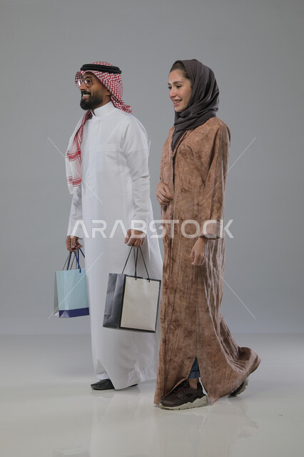 الاستفادة من العروض والتخفيضات الموسميه، تعابير وايماءات السرور والسعادة، شراء الهدايا والمستلزمات، بورتريه لزوجين عربيين خليجيين سعوديين يحملان بيديهما أكياس التسوق، الجسم بالطول الكامل، خلفية رمادية