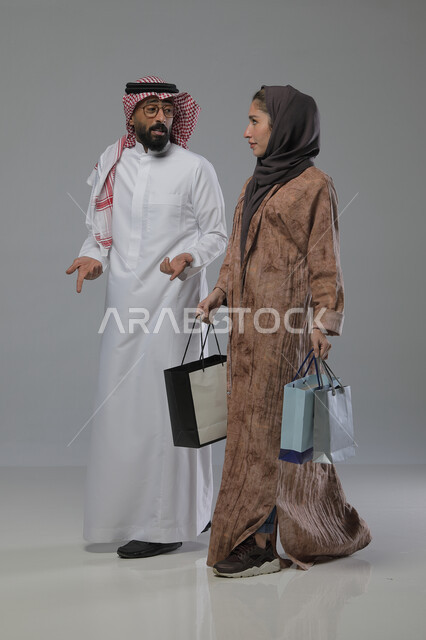 الاستفادة من العروض والتخفيضات الموسميه، تعابير وايماءات السرور والسعادة، شراء الهدايا والمستلزمات، بورتريه لزوجين عربيين خليجيين سعوديين يحملان بيديهما أكياس التسوق، الجسم بالطول الكامل، خلفية رمادية