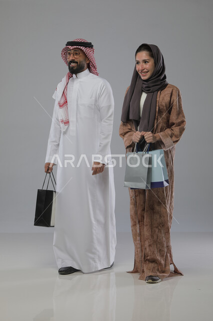 الاستفادة من العروض والتخفيضات الموسميه، تعابير وايماءات السرور والسعادة، شراء الهدايا والمستلزمات، بورتريه لزوجين عربيين خليجيين سعوديين يحملان بيديهما أكياس التسوق، الجسم بالطول الكامل، خلفية رمادية