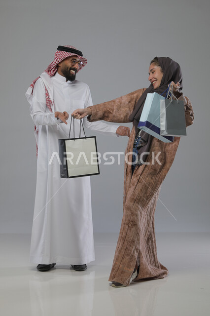 الاستفادة من العروض والتخفيضات الموسميه، تعابير وايماءات السرور والسعادة، شراء الهدايا والمستلزمات، بورتريه لزوجين عربيين خليجيين سعوديين يحملان بيديهما أكياس التسوق، الجسم بالطول الكامل، خلفية رمادية