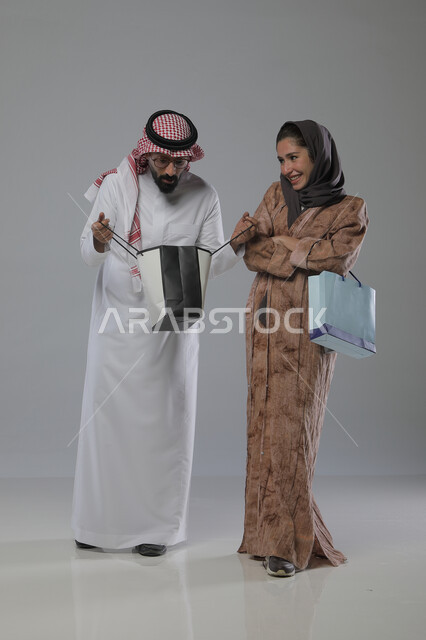 الاستفادة من العروض والتخفيضات الموسميه، تعابير وايماءات السرور والسعادة، شراء الهدايا والمستلزمات، بورتريه لزوجين عربيين خليجيين سعوديين يحملان بيديهما أكياس التسوق، الجسم بالطول الكامل، خلفية رمادية