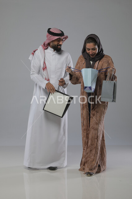 الاستفادة من العروض والتخفيضات الموسميه، تعابير وايماءات السرور والسعادة، شراء الهدايا والمستلزمات، بورتريه لزوجين عربيين خليجيين سعوديين يحملان بيديهما أكياس التسوق، الجسم بالطول الكامل، خلفية رمادية