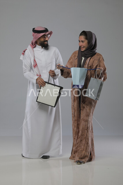 الاستفادة من العروض والتخفيضات الموسميه، تعابير وايماءات السرور والسعادة، شراء الهدايا والمستلزمات، بورتريه لزوجين عربيين خليجيين سعوديين يحملان بيديهما أكياس التسوق، الجسم بالطول الكامل، خلفية رمادية