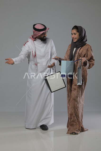 Taking advantage of seasonal offers and discounts, expressions and gestures of pleasure and happiness, buying gifts and supplies, portrait of a Saudi Arabian Gulf couple holding shopping bags in their hands, full body, gray background