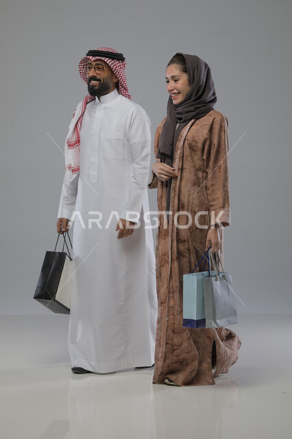 الاستفادة من العروض والتخفيضات الموسميه، تعابير وايماءات السرور والسعادة، شراء الهدايا والمستلزمات، بورتريه لزوجين عربيين خليجيين سعوديين يحملان بيديهما أكياس التسوق، الجسم بالطول الكامل، خلفية رمادية