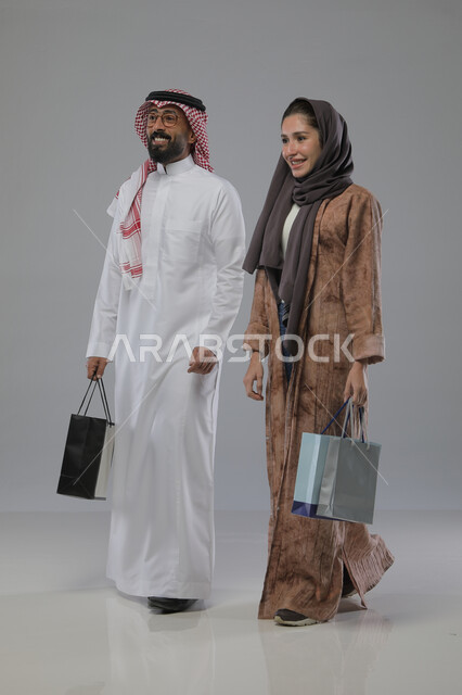 الاستفادة من العروض والتخفيضات الموسميه، تعابير وايماءات السرور والسعادة، شراء الهدايا والمستلزمات، بورتريه لزوجين عربيين خليجيين سعوديين يحملان بيديهما أكياس التسوق، الجسم بالطول الكامل، خلفية رمادية