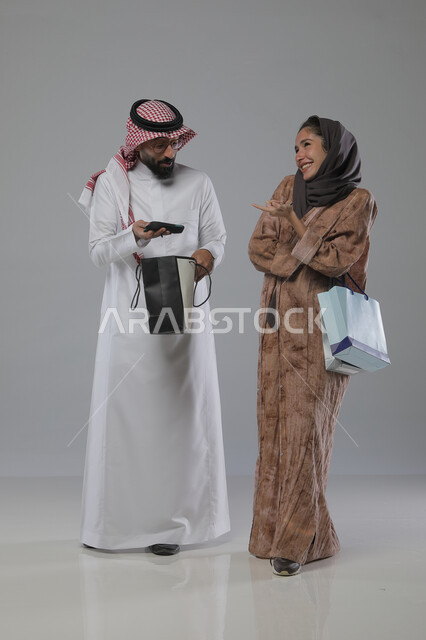 Taking advantage of seasonal offers and discounts, expressions and gestures of pleasure and happiness, buying gifts and supplies, portrait of a Saudi Arabian Gulf couple holding shopping bags in their hands, full body, gray background