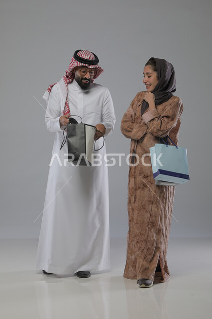 الاستفادة من العروض والتخفيضات الموسميه، تعابير وايماءات السرور والسعادة، شراء الهدايا والمستلزمات، بورتريه لزوجين عربيين خليجيين سعوديين يحملان بيديهما أكياس التسوق، الجسم بالطول الكامل، خلفية رمادية