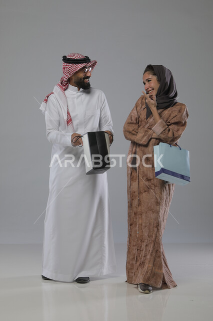 الاستفادة من العروض والتخفيضات الموسميه، تعابير وايماءات السرور والسعادة، شراء الهدايا والمستلزمات، بورتريه لزوجين عربيين خليجيين سعوديين يحملان بيديهما أكياس التسوق، الجسم بالطول الكامل، خلفية رمادية