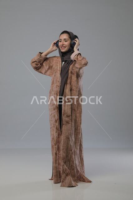 الاستماع الى الاغاني والموسيقى، استخدام جهاز تقني حديث، بورتريه لإمرأة عربية خليجية سعودية ترتدي الحجاب والعباءة تضع سماعات الهيدفون، تعابير السرور والسعادة، صورة الجسم بالطول الكامل، خلفية رمادية