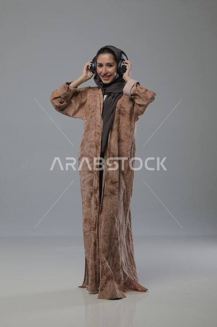 الاستماع الى الاغاني والموسيقى، استخدام جهاز تقني حديث، بورتريه لإمرأة عربية خليجية سعودية ترتدي الحجاب والعباءة تضع سماعات الهيدفون، تعابير السرور والسعادة، صورة الجسم بالطول الكامل، خلفية رمادية