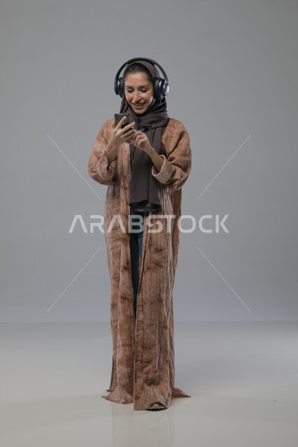  استخدام جهاز تقني حديث، الاستماع الى الاغاني والموسيقى، بورتريه لإمرأة عربية خليجية سعودية ترتدي الحجاب والعباءة تضع سماعات الهيدفون تمسك بيدها الهاتف المحمول، تعابير السرور والسعادة، صورة الجسم بالطول الكامل، خلفية رمادية