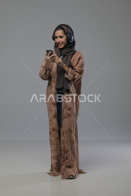  استخدام جهاز تقني حديث، الاستماع الى الاغاني والموسيقى، بورتريه لإمرأة عربية خليجية سعودية ترتدي الحجاب والعباءة تضع سماعات الهيدفون تمسك بيدها الهاتف المحمول، تعابير السرور والسعادة، صورة الجسم بالطول الكامل، خلفية رمادية
