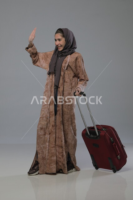 الشعور بالسعادة والفرح، الاستعداد لقضاء رحلة صيفية ترفيهية ممتعة, السياحة في إجازة الصيف, بورتريه لإمرأة عربية خليجية سعودية محجبة ترتدي العباءة تجر حقيبة السفر، إيماءات وتعابير الحماس, صورة الجسم بالطول الكامل، خلفية رمادية