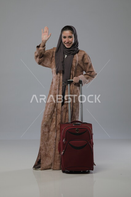 الشعور بالسعادة والفرح، الاستعداد لقضاء رحلة صيفية ترفيهية ممتعة, السياحة في إجازة الصيف, بورتريه لإمرأة عربية خليجية سعودية محجبة ترتدي العباءة تجر حقيبة السفر، إيماءات وتعابير الحماس, صورة الجسم بالطول الكامل، خلفية رمادية