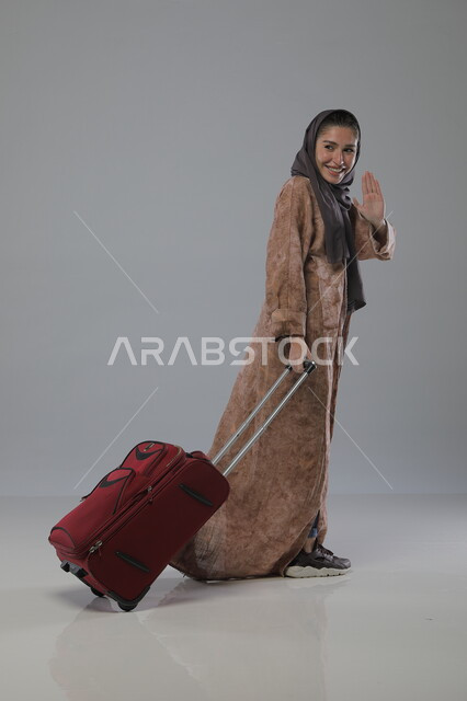 الشعور بالسعادة والفرح، الاستعداد لقضاء رحلة صيفية ترفيهية ممتعة, السياحة في إجازة الصيف, بورتريه لإمرأة عربية خليجية سعودية محجبة ترتدي العباءة تجر حقيبة السفر، إيماءات وتعابير الحماس, صورة الجسم بالطول الكامل، خلفية رمادية