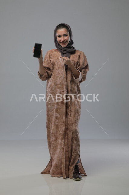 عرض الهاتف المحمول بشاشة سوداء فارغة، استخدام الجهاز التقني الحديث المتطور، بورتريه لامراة عربية خليجية سعودية محجبة ترتدي العباءة تنظر الى الكاميرا بايماءات السرور والسعادة، صورة الجسم بالطول الكامل، خلفية رمادية