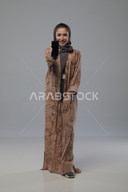 Mobile phone display with blank black screen, using modern advanced technical device, portrait of a veiled Saudi Arabian Gulf woman wearing abaya looking at the camera with gestures of pleasure and happiness, full body image, gray background