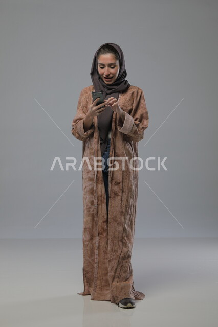 استخدام الجهاز التقني الحديث المتطور، بورتريه لامراة عربية خليجية سعودية محجبة ترتدي العباءة تعمل على الهاتف المحمول بايماءات السرور والاندماج، صورة الجسم بالطول الكامل، خلفية رمادية