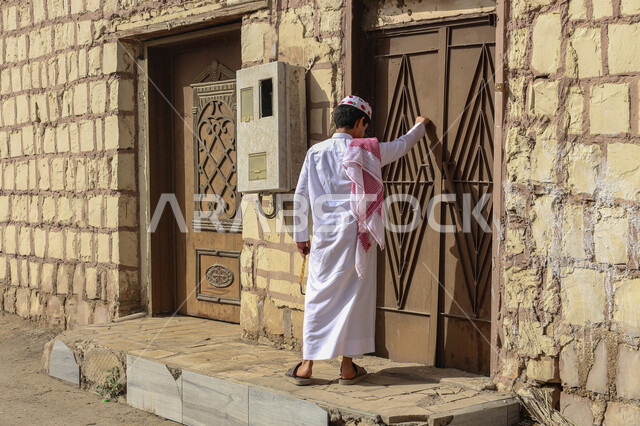 القرى والمدن التاريخية، التصميم المعماري العربي القديم للأبواب الأثرية القديمة، صورة من الخلف لولد عربي خليجي سعودي يرتدي الشماغ والثوب التقليدي يقوم بالدق على باب حديد لمنزل طيني، أماكن تراثية في المملكة العربية السعودية