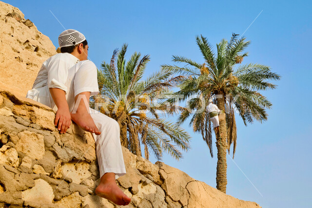 البيوت والمباني الطينية البسيطة، ولد عربي خليجي سعودي يرتدي زي تقليدي يجلس على صخرة جانب منزل من الطوب في إحدى الأحياء الشعبية في قرى المملكة العربية السعودية، الطراز العربي المعماري القديم، التأمل ومشاهدة المعالم والأماكن التاريخية التراثية المشهورة