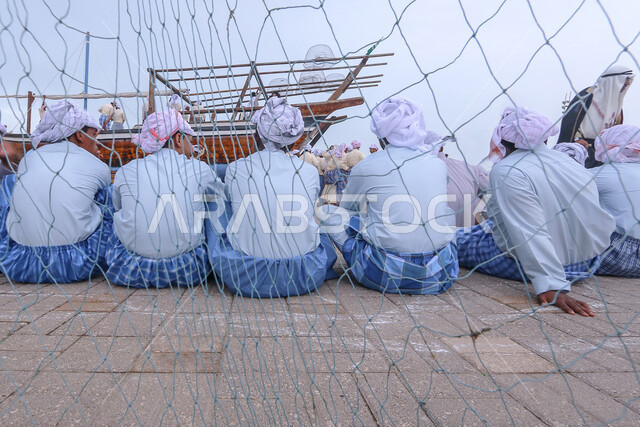 Practicing the profession of fishing and pearl diving in the Kingdom of Saudi Arabia, a photo from the back of a group of Saudi Arabian Gulf fishermen wearing traditional folk costume sitting in the middle of a wooden boat, a fishing net during the day