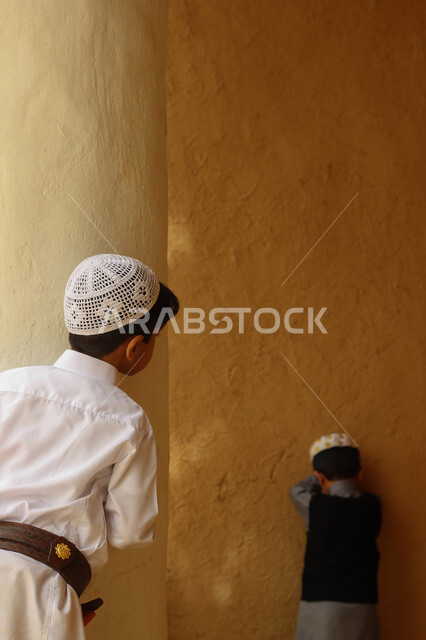 اللهو والمرح والاستمتاع بالوقت، ألعاب متوارثة عبر الأجيال، إحياء ذكرى تأسيس الدولة السعودية الأولى 1727 م, يوم بدينا 22 فبراير, منزل طيني قديم في قرية تاريخية أثرية، ولدان عربيان خليجيان سعوديان يرتديان الزي الشعبي التقليدي ويلعبان لعبة البحث والاختباء