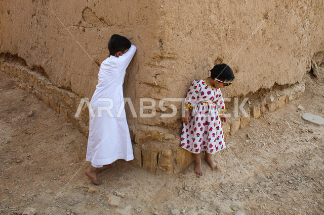 اللهو والمرح والاستمتاع بالوقت، ألعاب متوارثة عبر الأجيال، إحياء ذكرى تأسيس الدولة السعودية الأولى 1727 م, يوم بدينا 22 فبراير, منزل طيني قديم في قرية تاريخية أثرية، ولدان عربيان خليجيان سعوديان يرتديان الزي الشعبي التقليدي ويلعبان لعبة البحث والاختباء