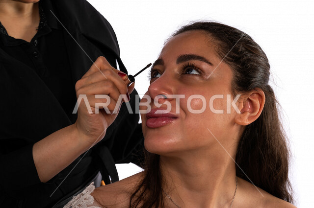 Portrait of a beautiful Saudi Arabian Gulf bride putting make-up by the hand of a make-up artist, using one of the cosmetics to decorate the bride, a wonderful photo session to prepare a Saudi bride, wearing a white wedding suit, the night of life, occasi