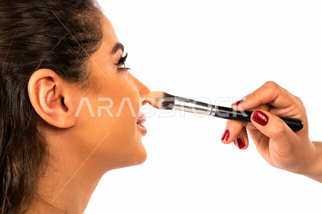 Portrait of a beautiful Saudi Arabian Gulf bride putting make-up by the hand of a make-up artist, using one of the cosmetics to decorate the bride, a wonderful photo session to prepare a Saudi bride, wearing a white wedding suit, the night of life, occasi