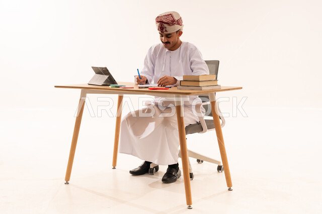 مفهوم التعلم عن بعد, البحث الالكتروني الذكي, بورتريه لطالب جامعي عربي خليجي عماني يرتدي الدشداشه والكمه يجلس على الطاولة ويعمل على الجهاز اللوحي, اسخدام الأجهزة والتقنيات المتطورة في المجال التعليمي , المناهج والمواد التعليميه في عمان, خلفية بيضاء