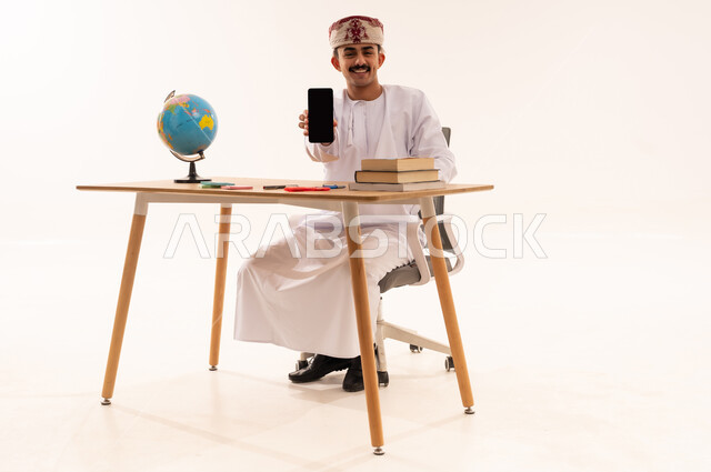 Academic education in Oman, displaying a blank black screen via mobile, academic universities and colleges, portrait of an Arab Gulf Omani university student wearing a keffiyeh and a dishdasha, smiling, sitting on a table holding a mobile phone in his hand, white background