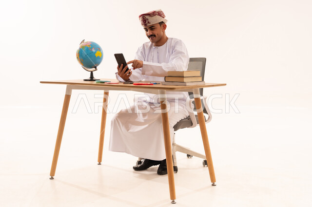 استخدام الأجهزة التقنية الحديثة , تصفح وسائل التواصل الاجتماعي عبر الجوال, بورتريه لطالب جامعي عربي خليجي عماني يرتدي الكمه والدشداشه مبتسم يجلس على الطاولة يمسك الهاتف المحمول بيده, خلفية بيضاء