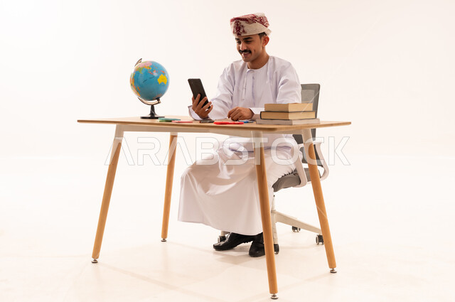 الإستعداد للمحاضرات الالكترونية،إستخدام أجهزة تقنية حديثة، بورتريه لطالب جامعي عربي خليجي عماني يرتدي الكمه والدشداشه مبتسم يجلس على الطاولة يقوم بإجراء مكالمة فيديو من خلال جهاز الهاتف المحمول، التعلم عن بعد ، مراجعة الدروس اليومية، خلفية بيضاء