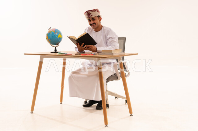 Enjoying reading, spending free time with useful activities, the concept of learning, excellence and diligence, curricula and educational materials in the Sultanate of Oman, portrait of a smiling Arab Gulf Omani university student wearing a keffiyeh and a dishdasha sitting at a table reading a book, white background