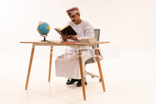 Spending free time with useful activities, the concept of learning, excellence and diligence, curricula and educational materials in the Sultanate of Oman, enjoying the hobby of reading, a portrait of an Arab Gulf Omani university student wearing a kemeh and a dishdasha sitting at a table reading a book, white background