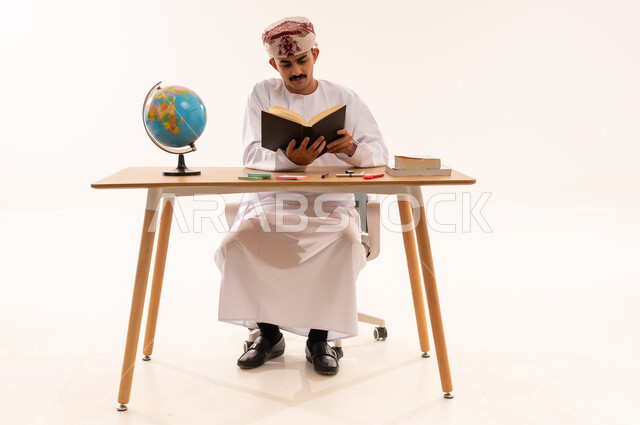 المناهج والمواد التعليمية في سلطنة عمان، مفهوم التعلم والتفوق والاجتهاد، قضاء أوقات الفراغ بالأنشطة المفيدة, الاستمتاع بممارسة هواية المطالعة, بورتريه لطالب جامعي عربي خليجي عماني يرتدي الكمه والدشداشه يجلس على الطاولة يقوم بقراءة الكتاب, خلفية بيضاء