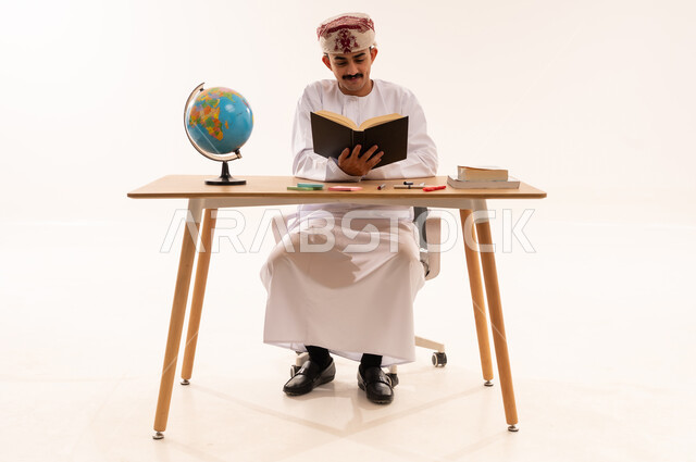 قضاء أوقات الفراغ بالأنشطة المفيدة, مفهوم التعلم والتفوق والاجتهاد, المناهج والمواد التعليمية في سلطنة عمان, الاستمتاع بممارسة هواية المطالعة, بورتريه لطالب جامعي عربي خليجي عماني يرتدي الكمه والدشداشه يجلس على الطاولة يقوم بقراءة الكتاب, خلفية بيضاء