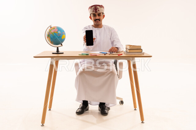 Blank black screen view on mobile, universities and colleges academic, portrait of an Arab Gulf Omani university student wearing a keffiyeh and a dishdasha smiling sitting on a table holding a mobile phone in his hand, academic education in Oman, full body, white background