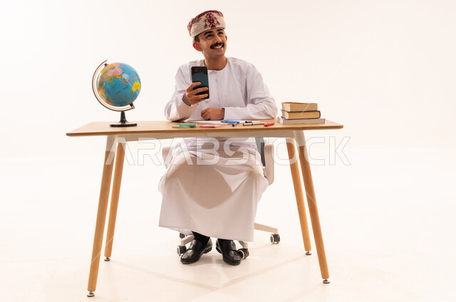 التعلم عن بعد ،الإستعداد للمحاضرات الالكترونية،إستخدام أجهزة تقنية حديثة، بورتريه لطالب جامعي عربي خليجي عماني يرتدي الكمه والدشداشه مبتسم يجلس على الطاولة يقوم بإجراء مكالمة فيديو من خلال جهاز الهاتف المحمول و ينظر الى شيء ما، مراجعة الدروس اليومية، الجسم بالطول الكامل، خلفية بيضاء