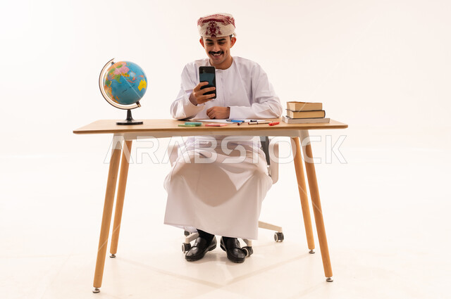 الإستعداد للمحاضرات الالكترونية،إستخدام أجهزة تقنية حديثة، بورتريه لطالب جامعي عربي خليجي عماني يرتدي الكمه والدشداشه مبتسم يجلس على الطاولة يقوم بإجراء مكالمة فيديو من خلال جهاز الهاتف المحمول، التعلم عن بعد ، مراجعة الدروس اليومية، الجسم بالطول الكامل، خلفية بيضاء