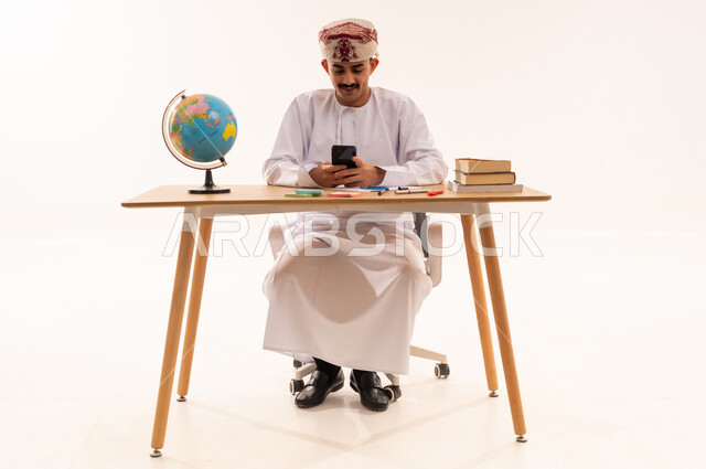 تصفح وسائل التواصل الاجتماعي عبر الجوال، استخدام الأجهزة التقنية الحديثة , بورتريه لطالب جامعي عربي خليجي عماني يرتدي الكمه والدشداشه يجلس على الطاولة يمسك الهاتف المحمول بيده , صورة للجسم بالطول الكامل, خلفية بيضاء