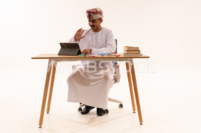 إستخدام أجهزة تقنية حديثة، الإستعداد للمحاضرات الالكترونية، بورتريه لطالب جامعي عربي خليجي عماني يرتدي الكمه والدشداشه يجلس على الطاولة يقوم بإجراء مكالمة فيديو من خلال جهاز التابلت ، التعلم عن بعد ، مراجعة الدروس اليومية، الجسم بالطول الكامل، خلفية بيضاء