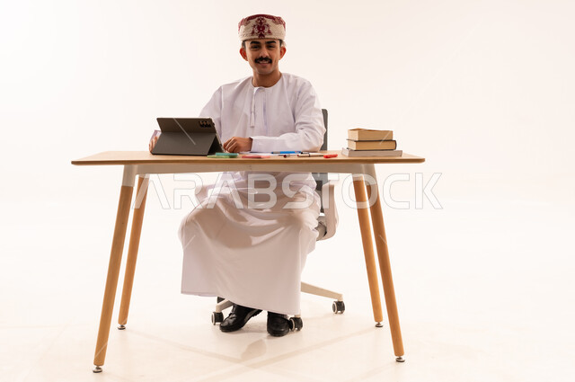البحث الالكتروني عبر الانترنت، استخدام الأجهزة والتقنيات الحديثة والمتطورة، مفهوم التعليم عن بعد, بورتريه لطالب جامعي عربي خليجي عماني يرتدي الكمه والدشداشه يجلس على الطاولة ويقوم بتدوين المعلومات على جهاز التابلت, مراجعة الدروس اليومية, خلفية بيضاء