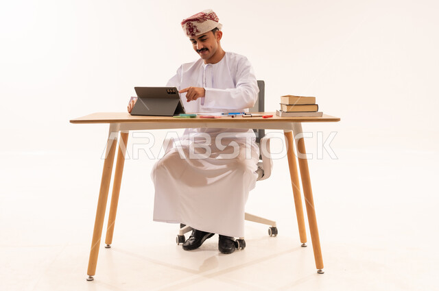 استخدام الأجهزة والتقنيات الحديثة والمتطورة، مفهوم التعليم عن بعد, بورتريه لطالب جامعي عربي خليجي عماني يرتدي الكمه والدشداشه يجلس على الطاولة ويقوم بتدوين المعلومات على جهاز التابلت, مراجعة الدروس اليومية, البحث الالكتروني عبر الانترنت, خلفية بيضاء