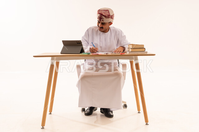 مراجعة الدروس اليومية، مفهوم التعليم عن بعد, بورتريه لطالب جامعي عربي خليجي عماني يرتدي الكمه والدشداشه يجلس على الطاولة ويقوم بتدوين المعلومات على جهازالتابلت, البحث الالكتروني عبر الانترنت, استخدام الأجهزة والتقنيات الحديثة والمتطورة, خلفية بيضاء