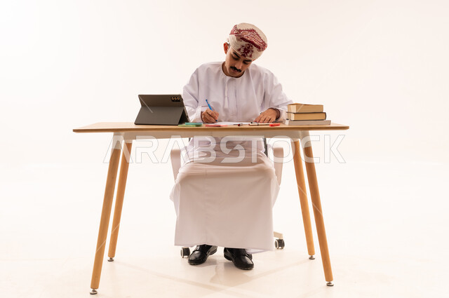 مفهوم التعليم عن بعد, بورتريه لطالب جامعي عربي خليجي عماني يرتدي الكمه والدشداشه يجلس على الطاولة ويقوم بتدوين المعلومات علي جهازالتابلت، مراجعة الدروس اليومية, البحث الالكتروني عبر الانترنت, استخدام الأجهزة والتقنيات الحديثة والمتطورة, خلفية بيضاء