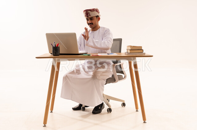 إستخدام أجهزه تقنية حديثة ، إيماءات تدل على السعادة ، بورتريه لطالب جامعي عربي خليجي عماني يرتدي الكمه والدشداشه يجلس على الطاولة يقوم بإجراء مكالمة فيديو من خلال كمبيوتر محمول ، الإستعداد للمحاضرات الجامعية الالكترونية ، التعلم عن بعد ، مراجعة الدروس اليومية ، خلفية زرقاء