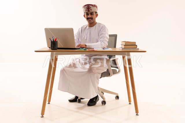مفهوم التعليم عن بعد، بورتريه لطالب جامعي عربي خليجي عماني يرتدي الكمه والدشداشه يجلس على الطاولة يستخدم  الحاسب المحمول في البحث والمطالعة وتصفح التطبيقات الالكترونية، ايماءات الوجه تدل على النجاح والامتياز، خلفية بيضاء