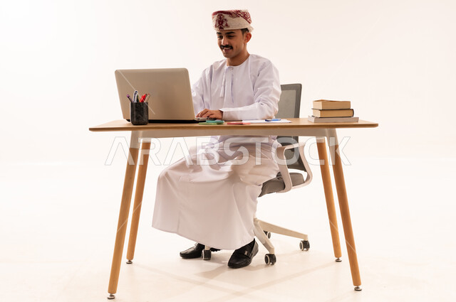 الاستعداد للعام الدراسي الجامعي الجديد، بورتريه لطالب جامعي عربي خليجي عماني يرتدي الكمه والدشداشه يجلس على الطاولة يستخدم  الحاسب المحمول في البحث والمطالعة وتصفح التطبيقات الالكترونية، ايماءات الوجه واليد تدل على النجاح والامتياز، خلفية بيضاء